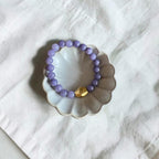 A beaded stretch bracelet with light purple round beads and a single gold accent bead, displayed on a small scalloped ceramic dish over a white fabric background.