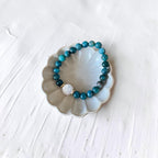 A beaded stretch bracelet with turquoise-blue and dark teal beads, featuring a white textured bead and a small gold spacer, displayed on a scalloped white ceramic dish over a white textured background.