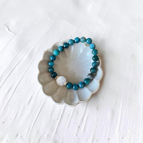 A beaded stretch bracelet with turquoise-blue and dark teal beads, featuring a white textured bead and a small gold spacer, displayed on a scalloped white ceramic dish over a white textured background.