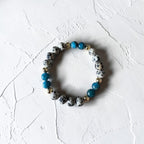 A beaded stretch bracelet featuring white and black spotted beads, bright turquoise blue beads, and small metallic gold spacer beads, displayed on a textured white surface.
