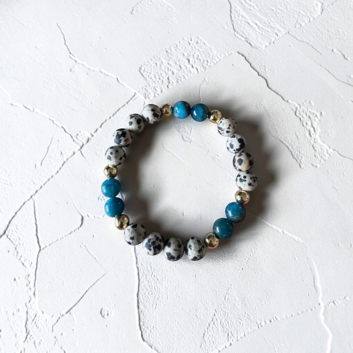 A beaded stretch bracelet featuring white and black spotted beads, bright turquoise blue beads, and small metallic gold spacer beads, displayed on a textured white surface.