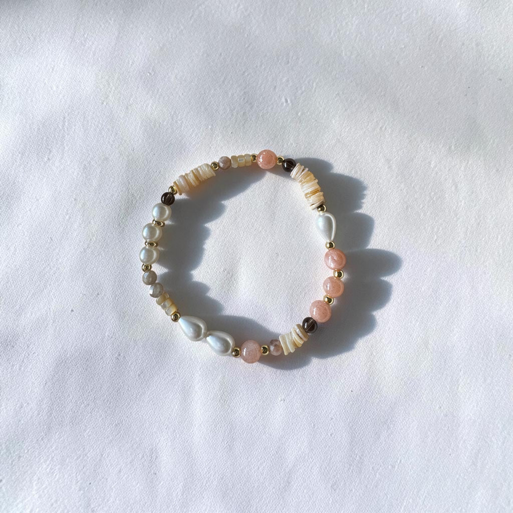 A beaded stretch bracelet featuring white pearls, cream striped beads, light pink beads, and small gold spacer beads, displayed on a textured white fabric background.