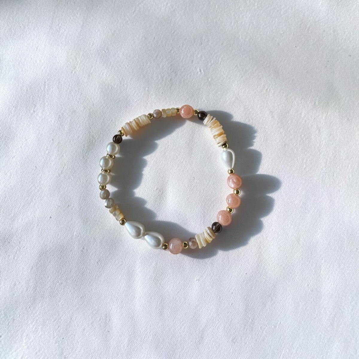 A beaded stretch bracelet featuring white pearls, cream striped beads, light pink beads, and small gold spacer beads, displayed on a textured white fabric background.