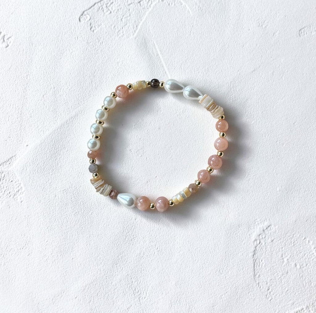 A delicate beaded stretch bracelet featuring pink, white, and gray beads with gold spacer accents, displayed on a textured white surface.