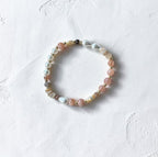 A delicate beaded stretch bracelet featuring pink, white, and gray beads with gold spacer accents, displayed on a textured white surface.