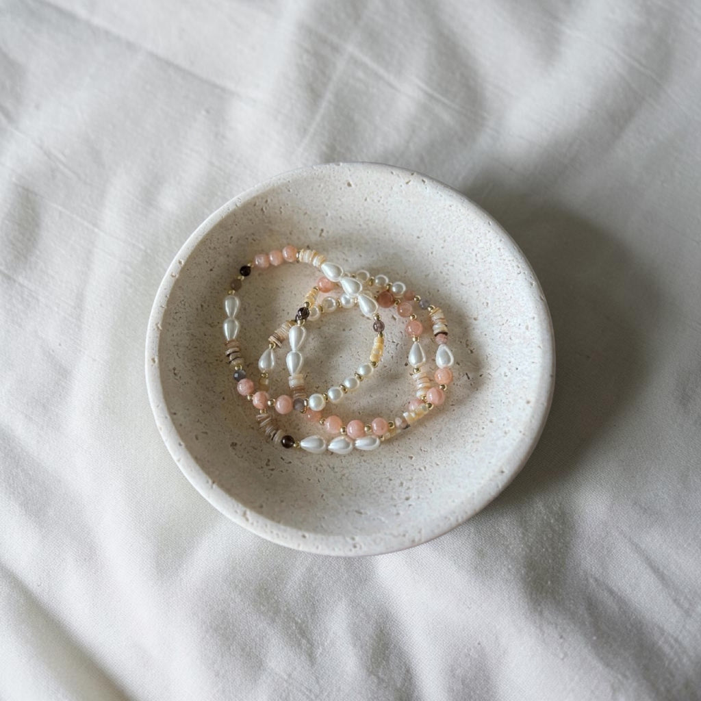 Three beaded stretch bracelets with white, pink, and gold-toned beads resting in a small speckled ceramic dish on a white fabric background.