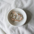 Three beaded stretch bracelets with white, pink, and gold-toned beads resting in a small speckled ceramic dish on a white fabric background.