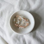 Three beaded stretch bracelets with white, pink, and gold-toned beads resting in a small speckled ceramic dish on a white fabric background.