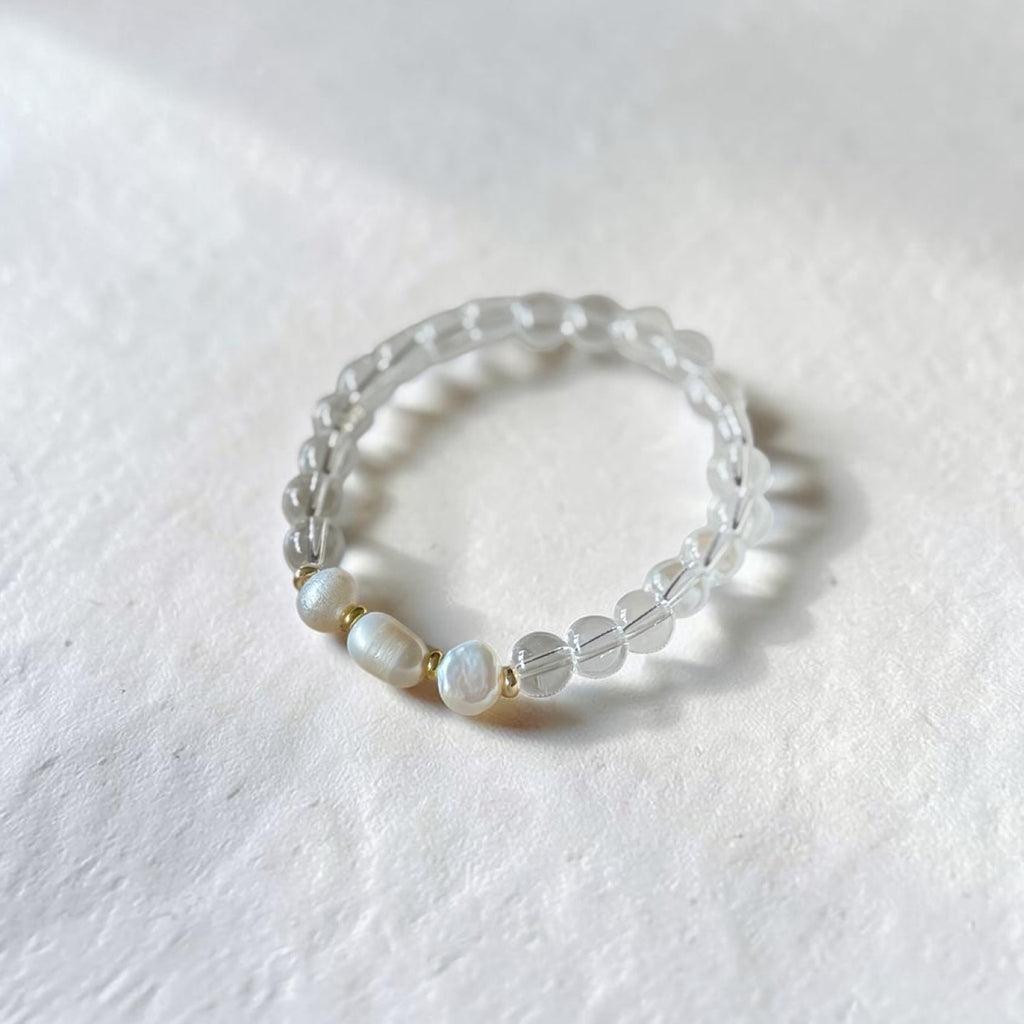 A beaded stretch bracelet featuring clear faceted beads and three white mother-of-pearl beads separated by small gold spacers, resting on a textured white surface.