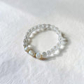 A beaded stretch bracelet featuring clear faceted beads and three white mother-of-pearl beads separated by small gold spacers, resting on a textured white surface.