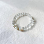 A beaded stretch bracelet featuring clear faceted beads and three white mother-of-pearl beads separated by small gold spacers, resting on a textured white surface.