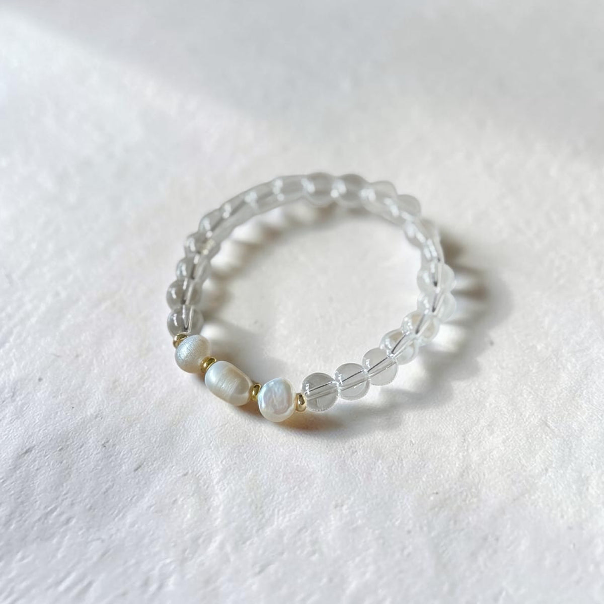 A beaded stretch bracelet featuring clear faceted beads and three white mother-of-pearl beads separated by small gold spacers, resting on a textured white surface.