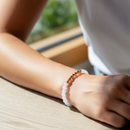 A close-up of a person's wrist wearing a beaded bracelet with white round beads and a small section of metallic gold beads, resting on a light wood table.