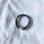 A beaded stretch bracelet featuring dark gray stone beads and a single white pearl accent bead, displayed on a textured white fabric background.