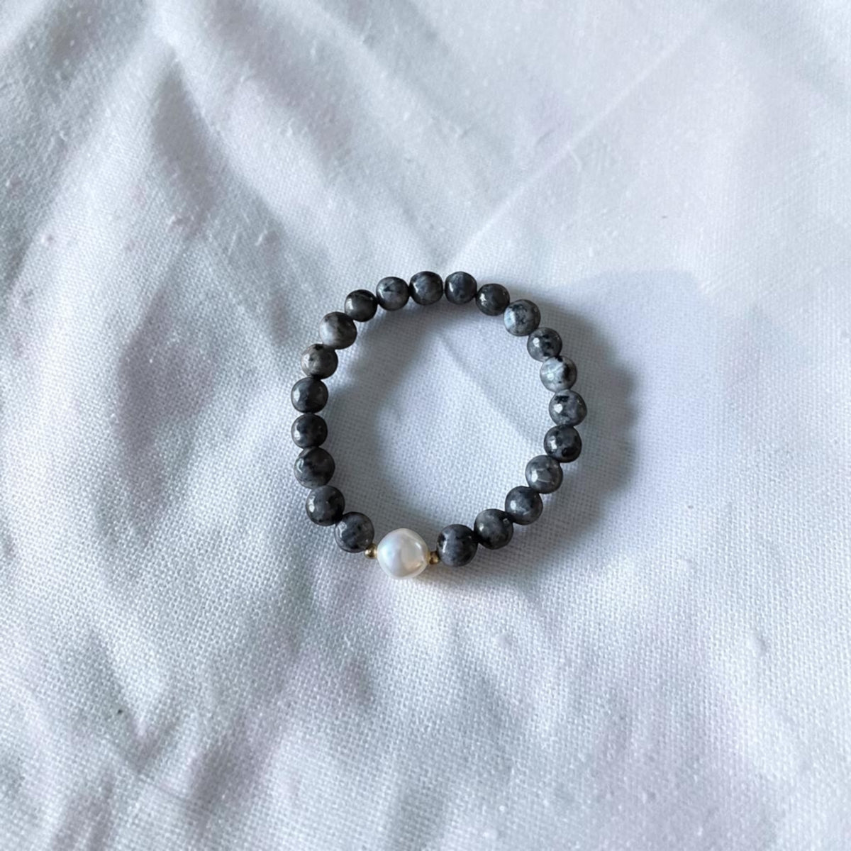A beaded stretch bracelet featuring dark gray stone beads and a single white pearl accent bead, displayed on a textured white fabric background.