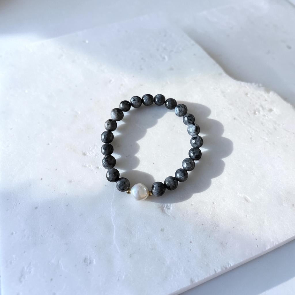 A beaded stretch bracelet made of dark gray to black round beads with one small white pearl accent and tiny gold spacer beads, displayed on a white stone tile.