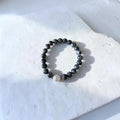 A beaded stretch bracelet made of dark gray to black round beads with one small white pearl accent and tiny gold spacer beads, displayed on a white stone tile.