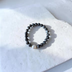 A beaded stretch bracelet made of dark gray to black round beads with one small white pearl accent and tiny gold spacer beads, displayed on a white stone tile.