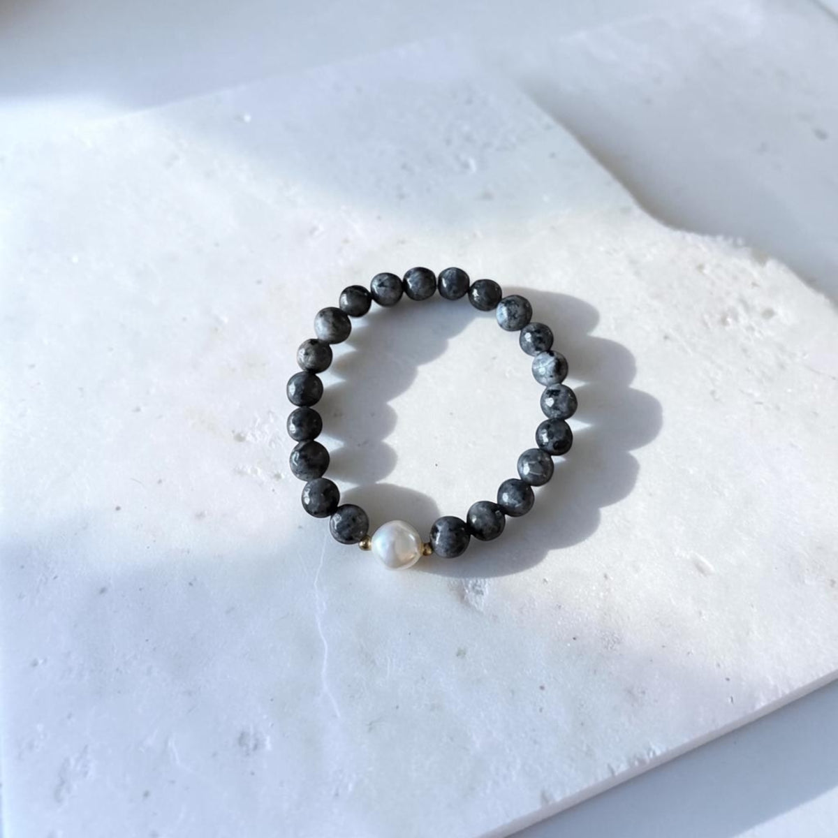 A beaded stretch bracelet made of dark gray to black round beads with one small white pearl accent and tiny gold spacer beads, displayed on a white stone tile.