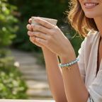 A woman outdoors wearing a light blue beaded bracelet with a small gold bow charm, holding a cup in her hands.