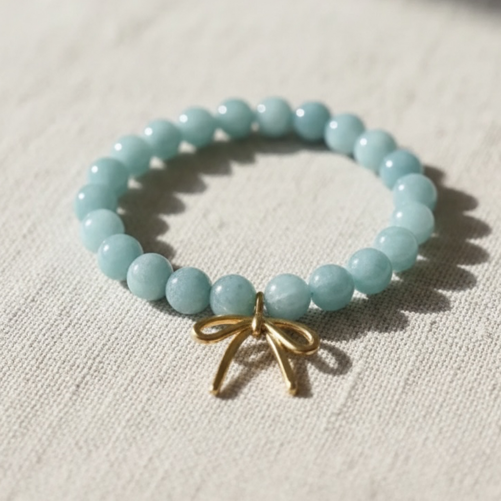A beaded stretch bracelet made of light aqua blue round beads with a small gold bow charm, resting on a textured beige linen fabric.