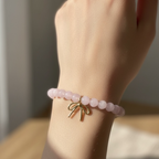 A close-up of a person's wrist wearing a beaded bracelet made of pale pink round beads with a small gold bow charm at the center, set against a softly blurred indoor background.