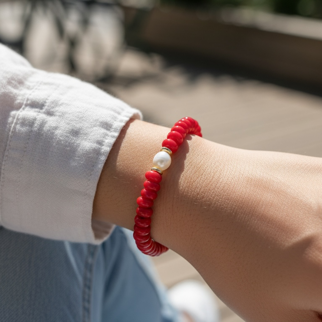 A close-up of a person's wrist wearing a red beaded bracelet with a white pearl and small gold spacer, photographed outdoors in bright natural light.