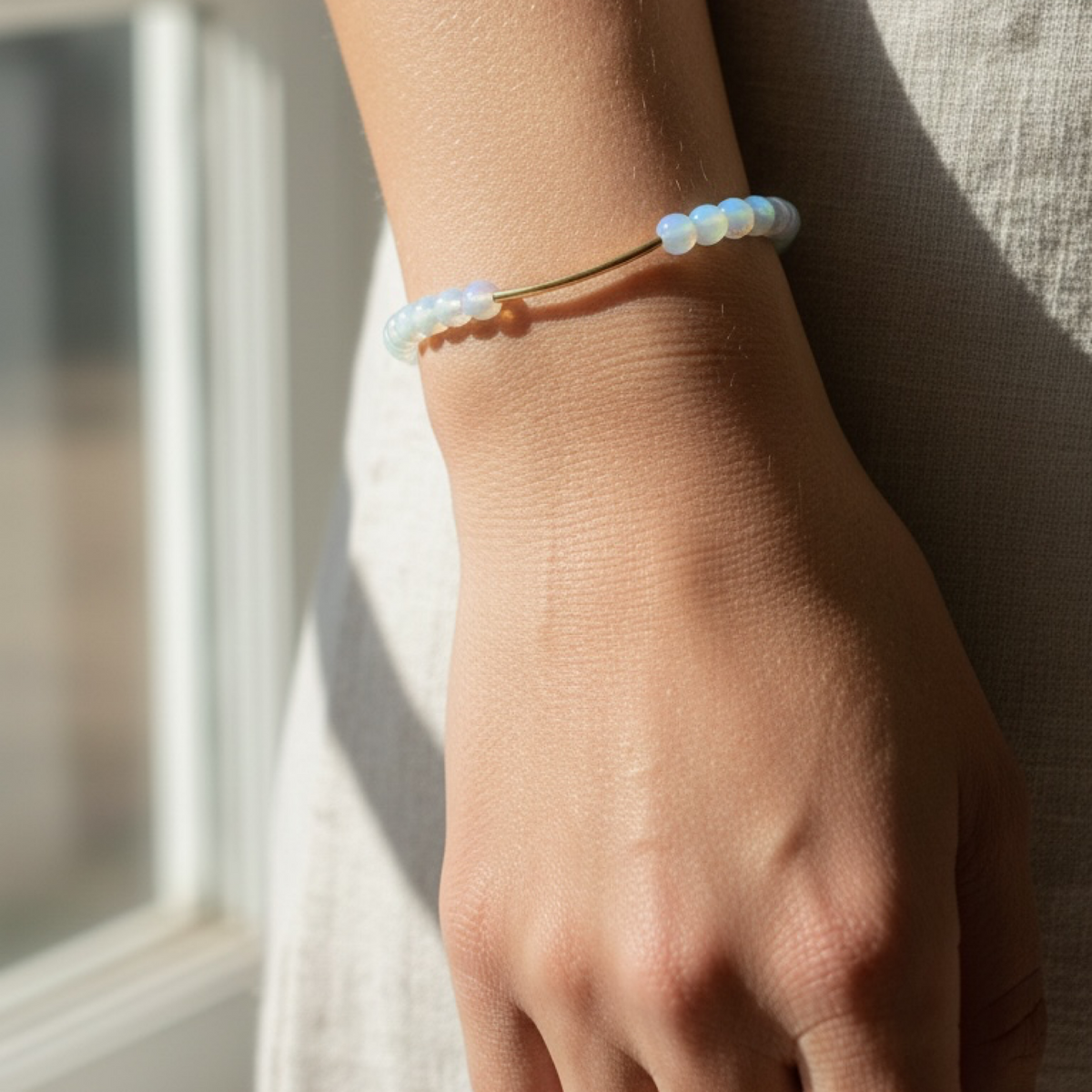 A delicate beaded bracelet with iridescent white and pale blue beads on a thin gold band, shown worn on a person's wrist in warm natural light.