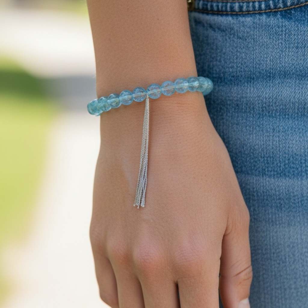 A close-up of a person's wrist wearing a light blue beaded bracelet with a dangling silver tassel, paired with blue denim jeans.