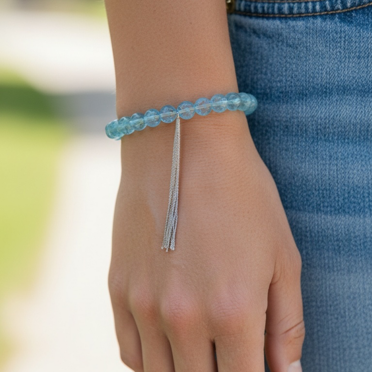 A close-up of a person's wrist wearing a light blue beaded bracelet with a dangling silver tassel, paired with blue denim jeans.