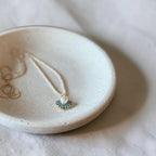 A small round ceramic jewelry tray holding a delicate necklace with a small blue and white pendant, set on a softly wrinkled white fabric background.