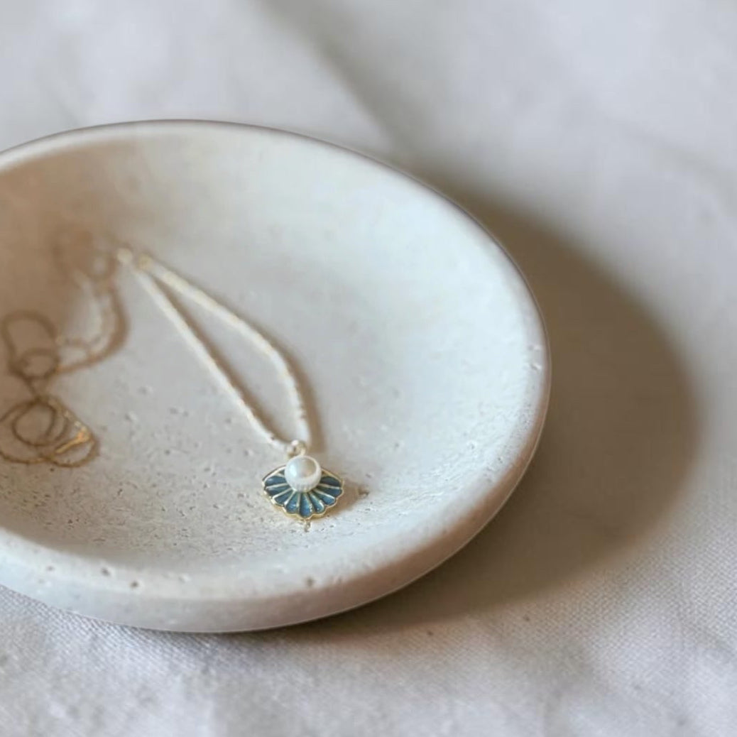 A small round ceramic jewelry tray holding a delicate necklace with a small blue and white pendant, set on a softly wrinkled white fabric background.