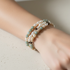 A close-up of a person's wrist wearing two beaded wrap bracelets, one in pale green and cream beads and the other in cream and white pearls, resting on a textured light beige stone block.