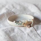 A delicate beaded bracelet with light blue, white, and gold beads rests inside a small, shallow off-white ceramic dish on a textured white fabric background.