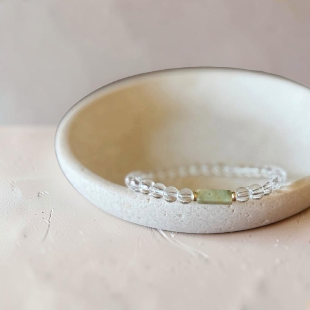 A beaded stretch bracelet with clear faceted beads and a central light green rectangular stone, displayed inside a small off-white ceramic dish.