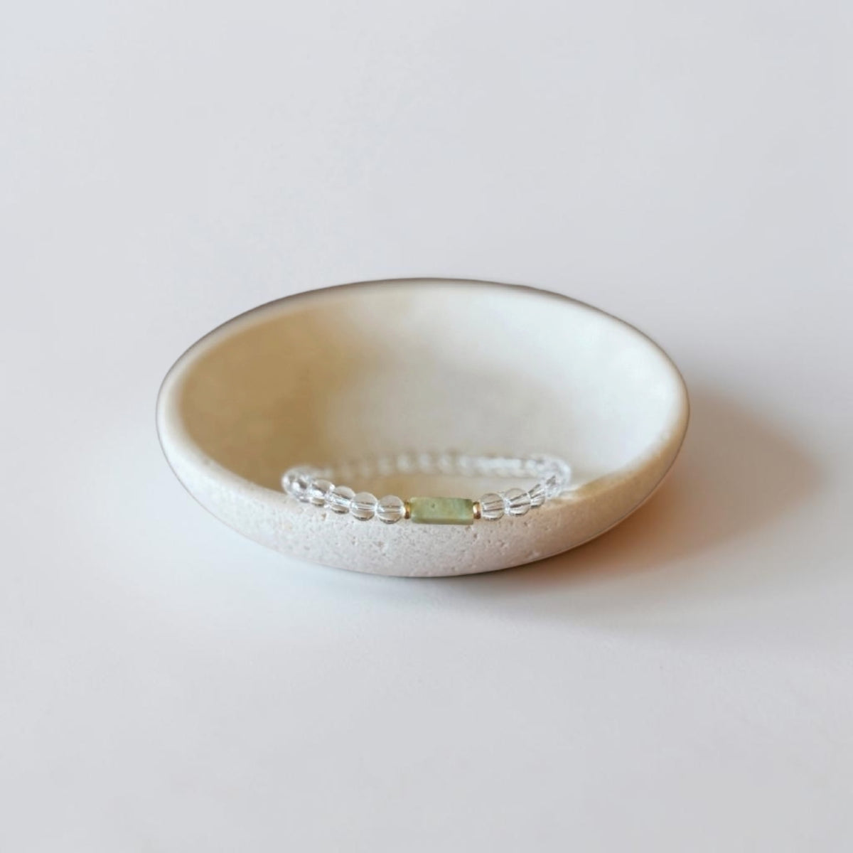 A delicate beaded bracelet with a small light green rectangular stone centerpiece, displayed inside a shallow, speckled cream ceramic dish on a soft, light gray background.