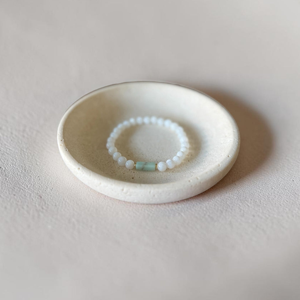 A delicate beaded bracelet with white round beads and a small mint green accent bead, displayed inside a small, round, textured ceramic dish on a neutral beige surface.
