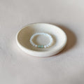A delicate beaded bracelet with white round beads and a small mint green accent bead, displayed inside a small, round, textured ceramic dish on a neutral beige surface.