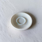 A delicate beaded bracelet with white round beads and a small mint green accent bead, displayed inside a small, textured off-white ceramic dish on a neutral concrete surface.