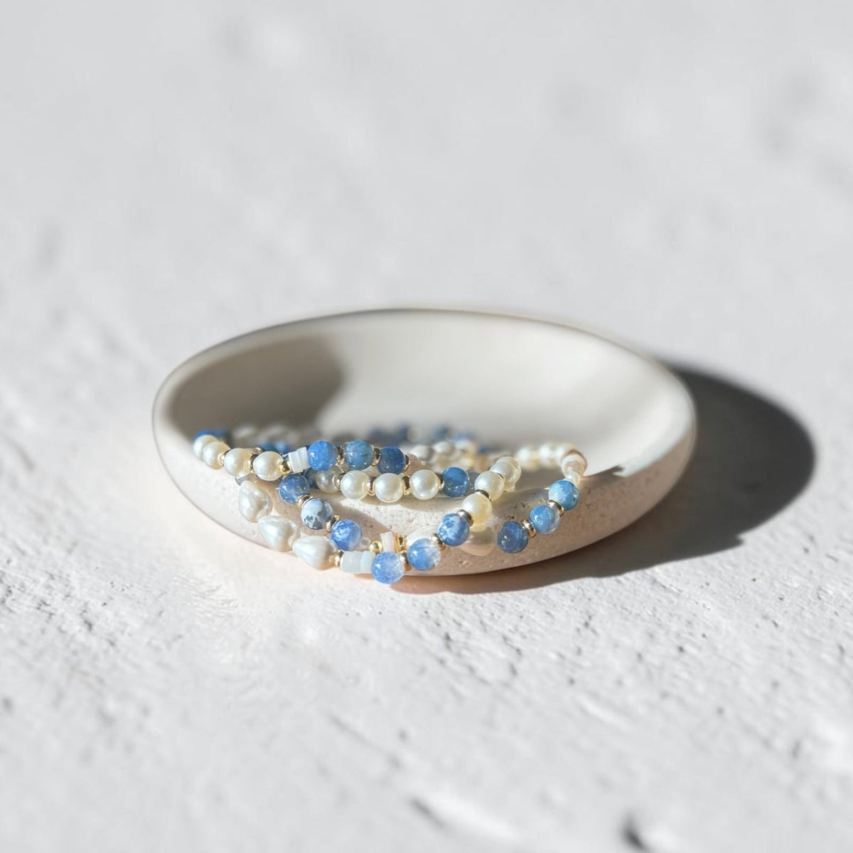 A set of delicate beaded strands in light blue and cream-white, arranged in a small white ceramic dish on a textured white surface.