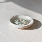A small round white ceramic dish holding several beaded stretch bracelets in light green, white, and metallic tones, photographed on a sunlit off-white surface with a soft shadow.