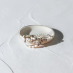 A delicate beaded strand bracelet with white pearls, brown beads, and small iridescent rhinestone accents, draped over a small white ceramic dish on a textured white surface.