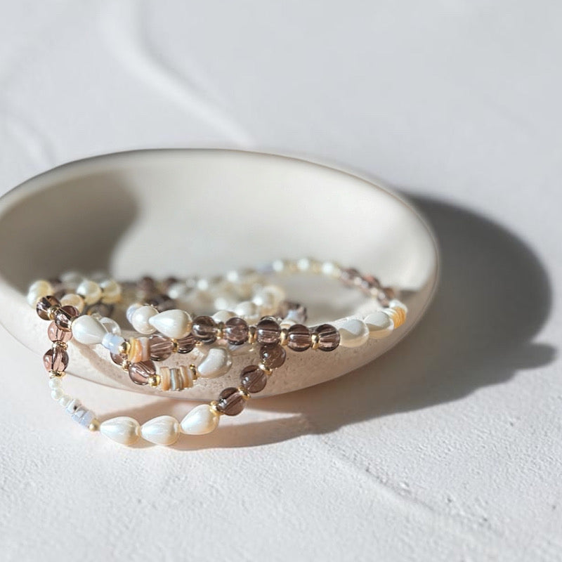 A beaded strand jewelry piece with white pearl-like beads, brown translucent beads, and small striped spacer beads, draped over a small round ceramic dish on a light textured surface.