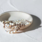 A beaded strand jewelry piece with white pearl-like beads, brown translucent beads, and small striped spacer beads, draped over a small round ceramic dish on a light textured surface.