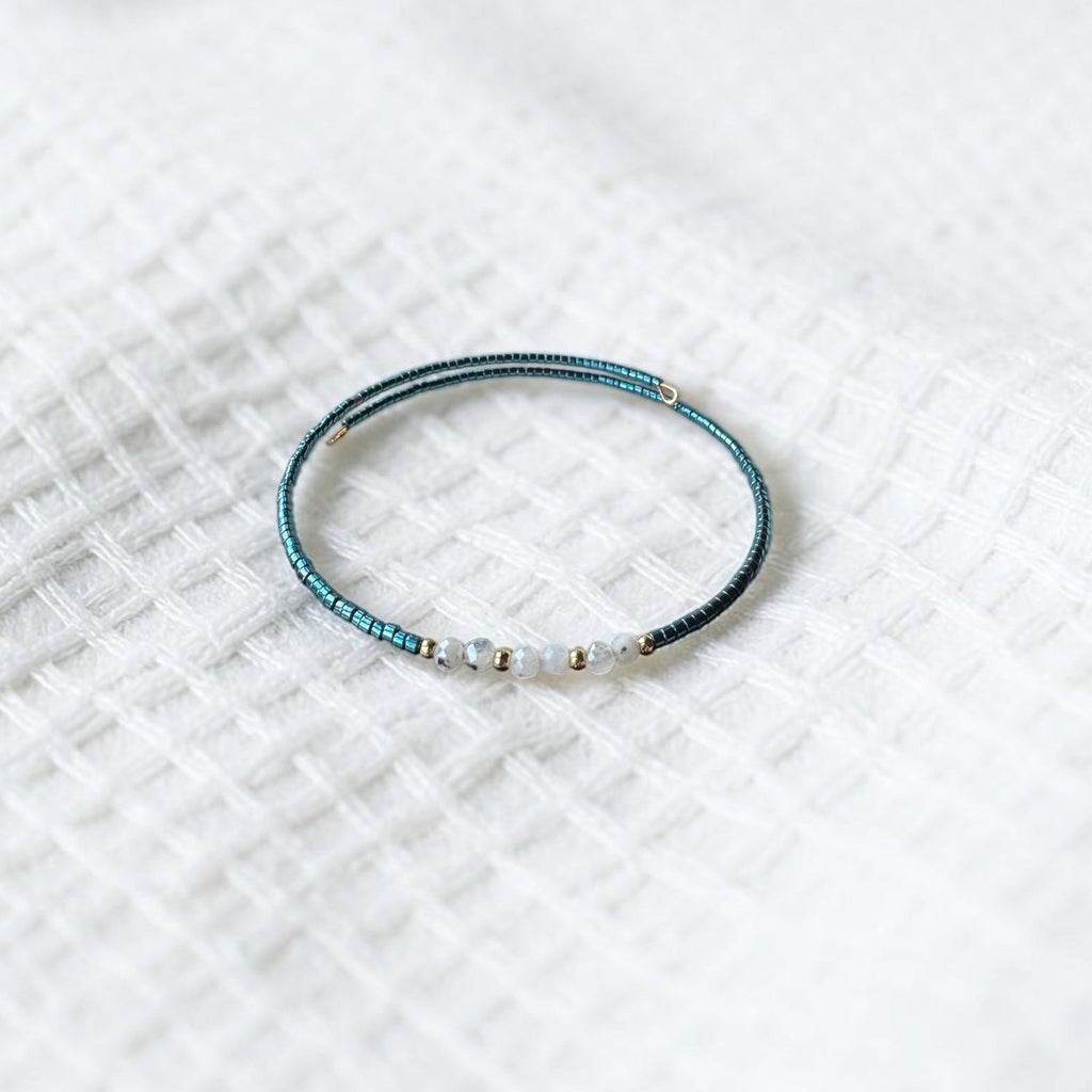 A delicate beaded bracelet with a dark teal-blue textured body and small white beads, resting on a white textured fabric background.