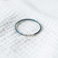 A delicate beaded bracelet with a dark teal-blue textured body and small white beads, resting on a white textured fabric background.