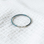 A delicate beaded bracelet with a dark teal-blue textured body and small white beads, resting on a white textured fabric background.
