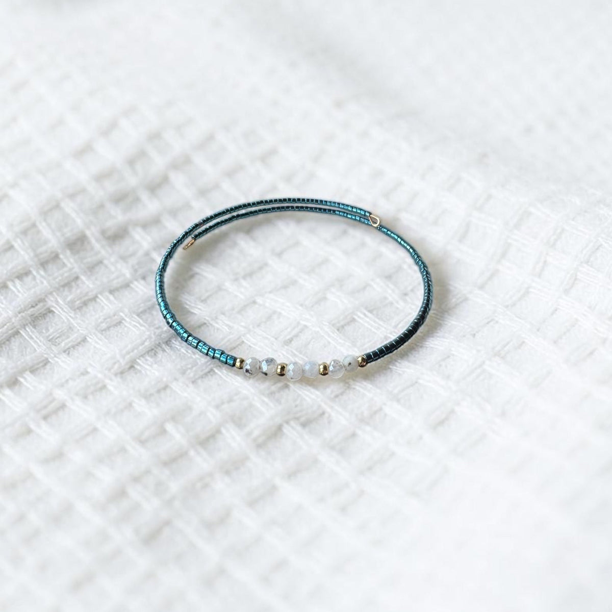 A delicate beaded bracelet with a dark teal-blue textured body and small white beads, resting on a white textured fabric background.