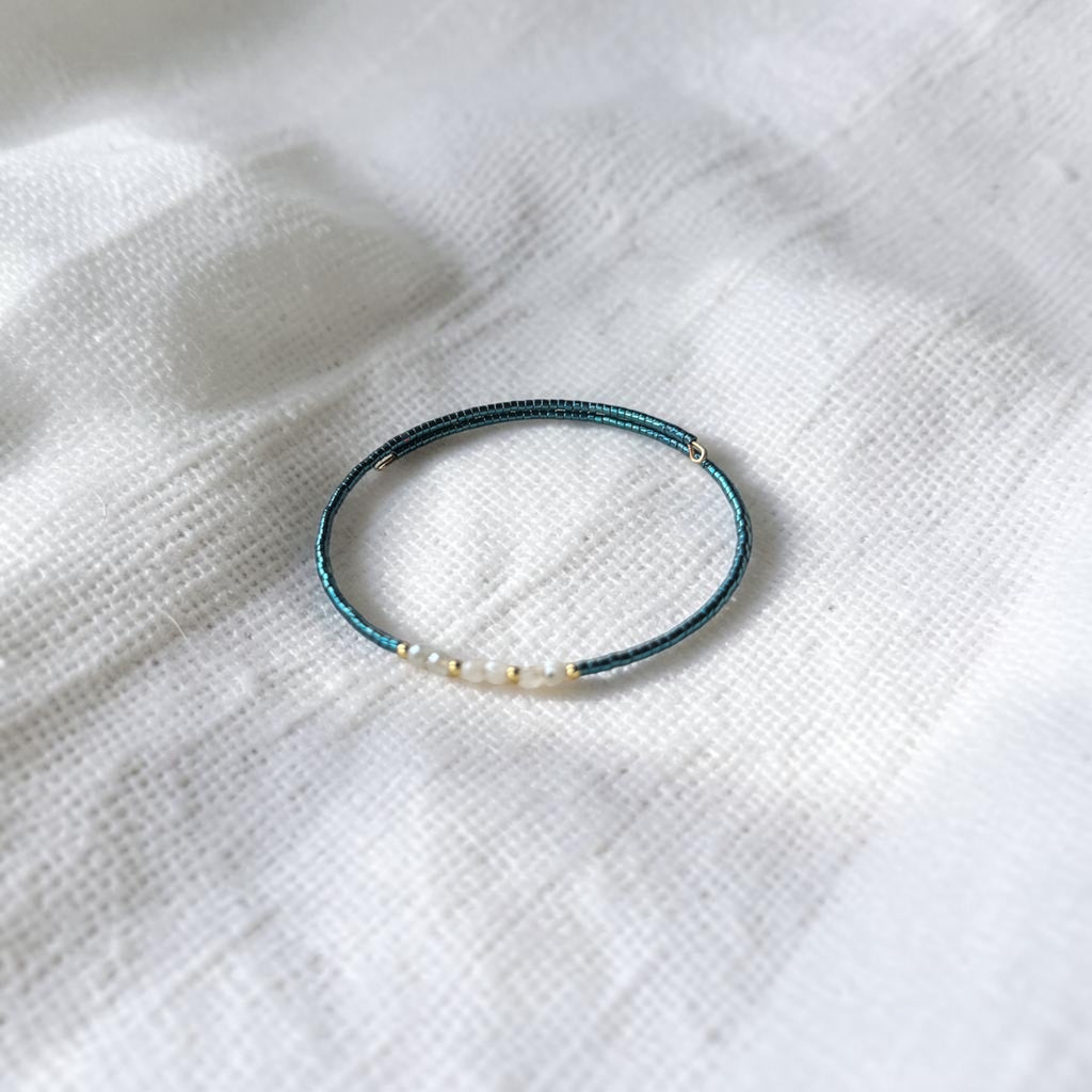 A delicate beaded bracelet with a dark teal-blue wrap, accented by small white pearls and metallic gold beads, resting on a textured white fabric background.