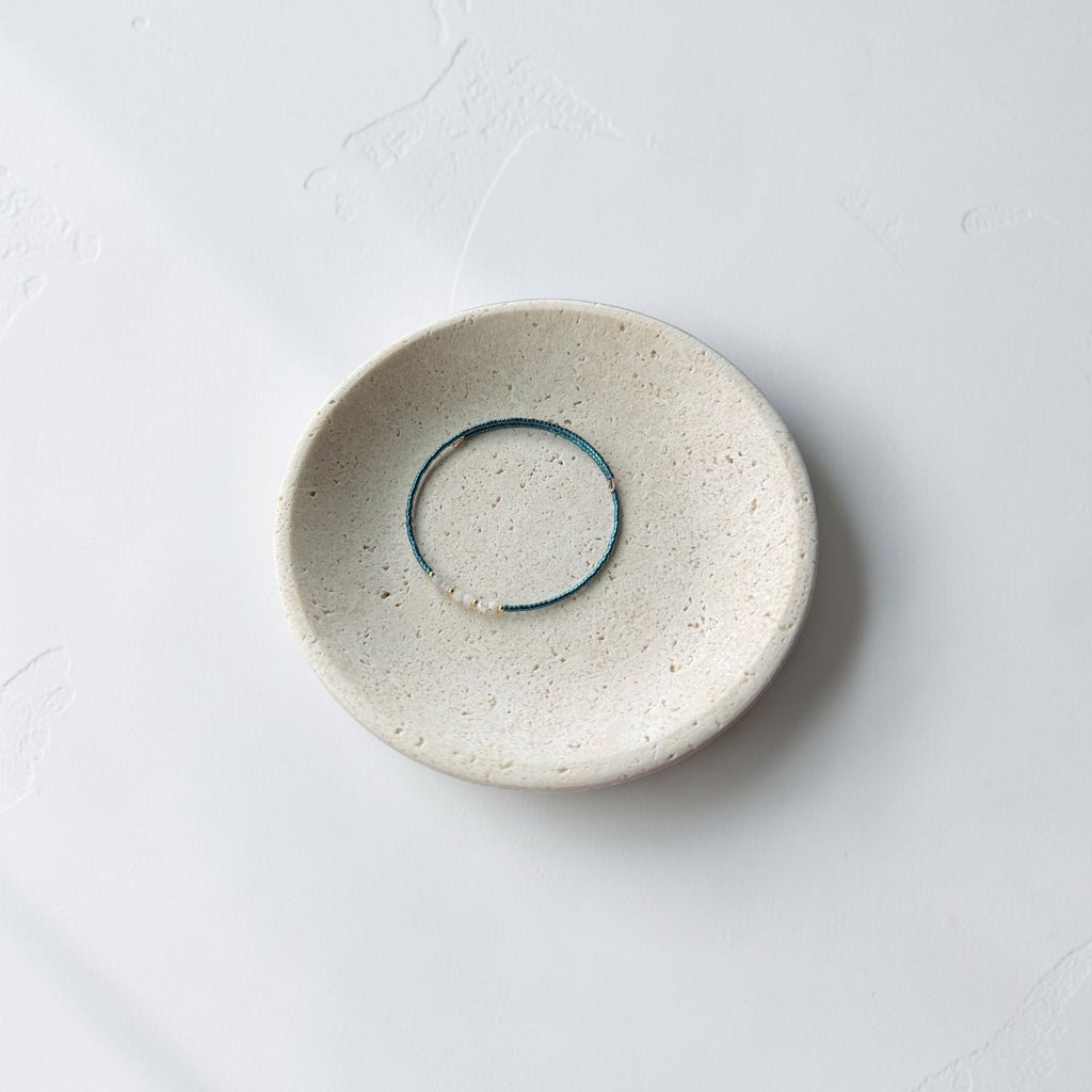 A minimalist ceramic dish with a textured cream-beige glaze, featuring a thin blue teal wire bracelet inside its shallow center, set against a clean white background.